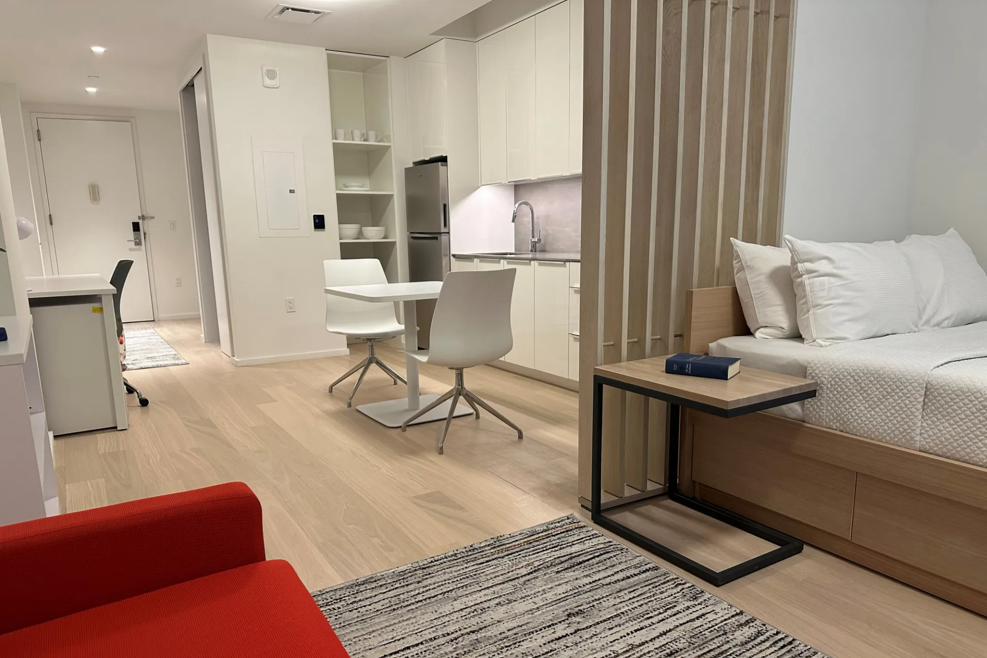 A furnished studio apartment in 600 W. 125th Street, with white cabinetry, a wood accent wall separating the kitchen and the bed.