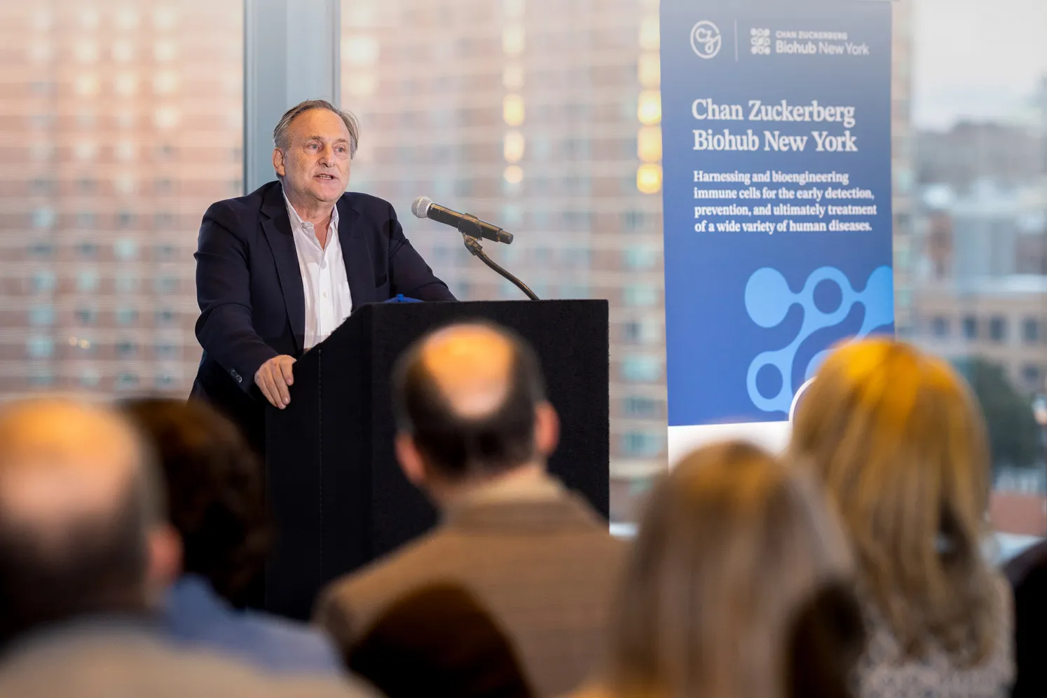 Dr. Andrea Califano talks from a podium in front of a group of people. Next to him, a banner reads, "Chan Zuckerberg Biohub New York - harnessing and bioengineering immune cells for the early detection, prevention, and ultimately treatment of a wide variety of human diseases."