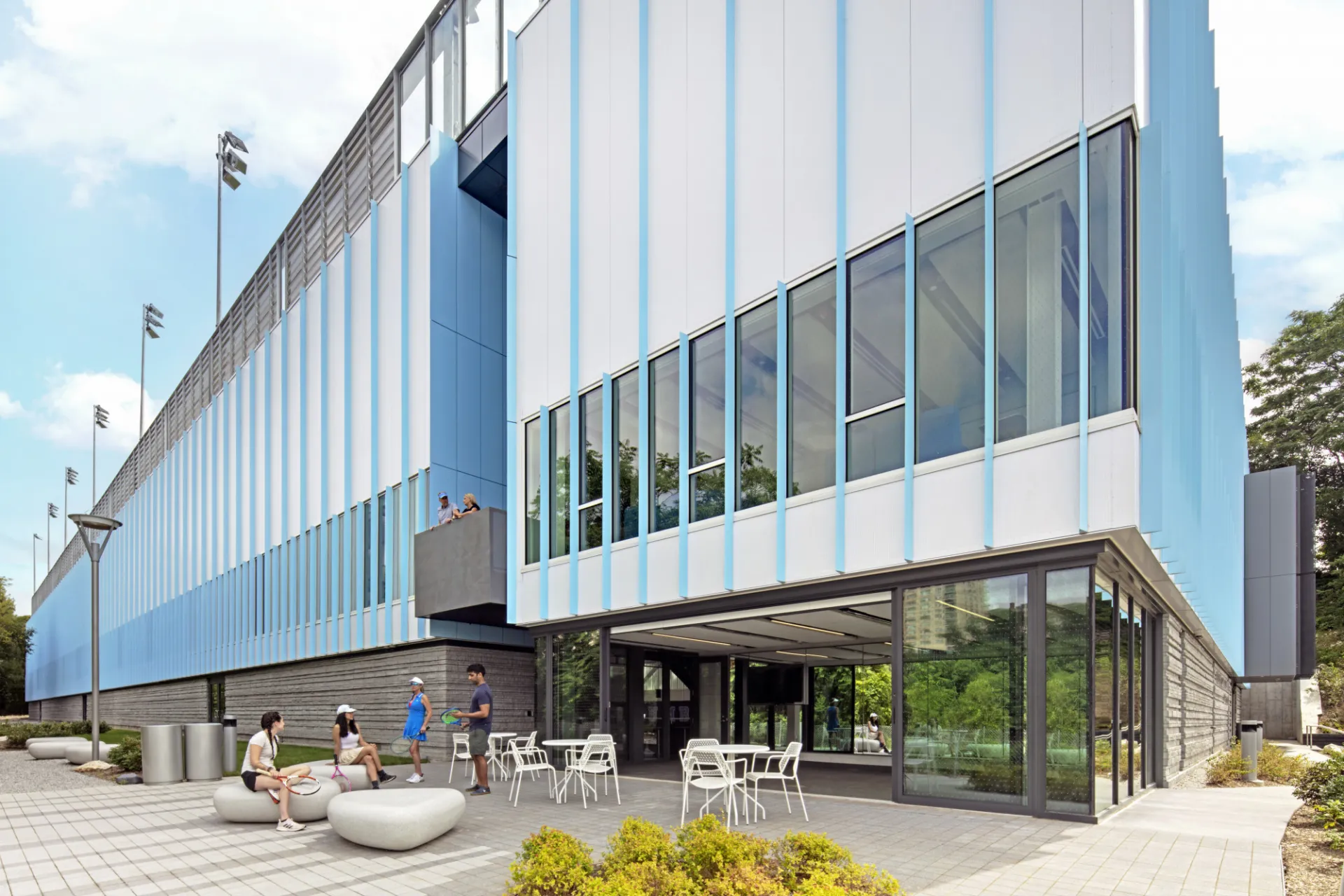 The exterior of the Milstein Tennis Center, which has white facade panels with light blue accents, and stadium lighting on the roof.