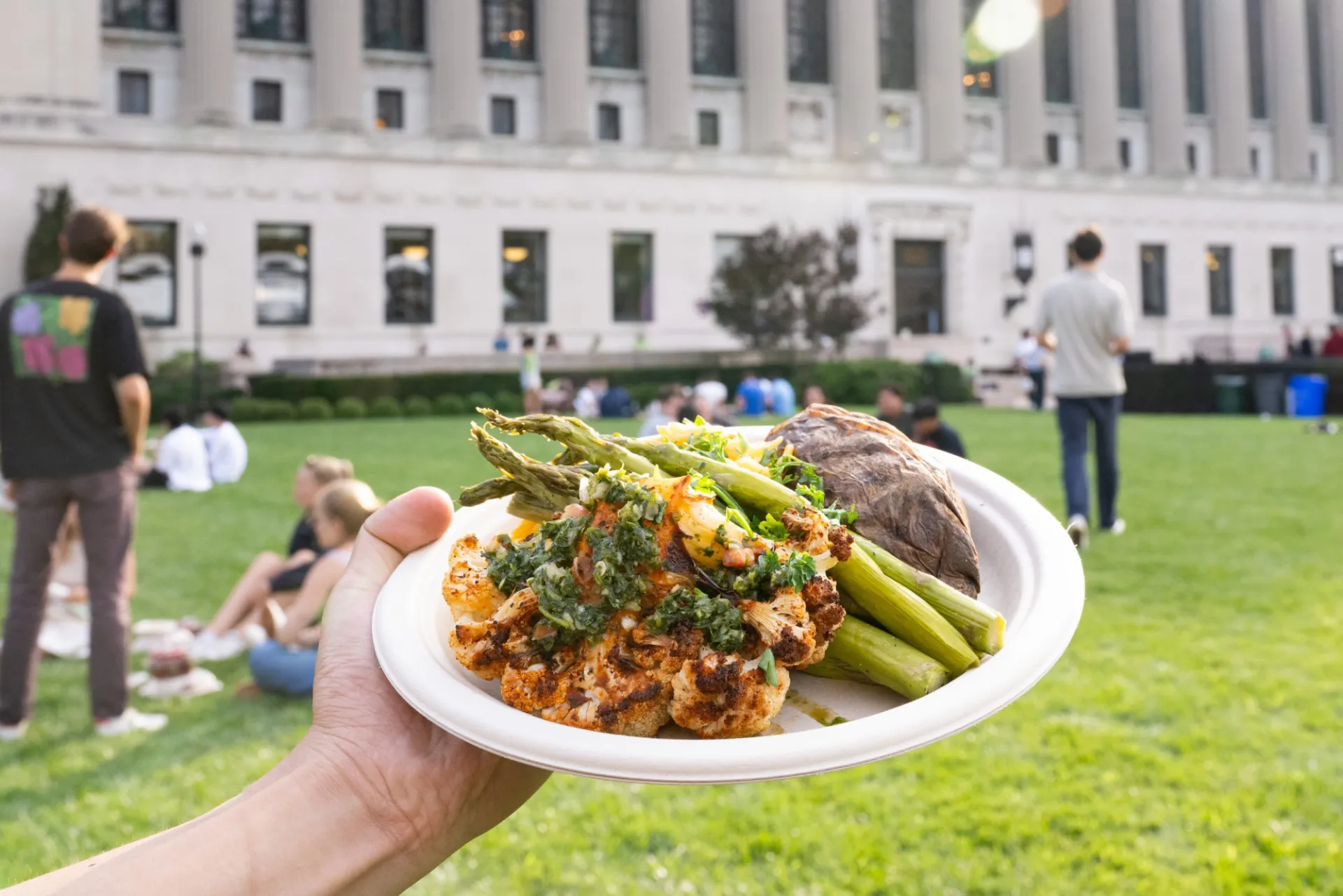 A cauliflower steak topped with chimichurri sauce on a compostable plate. Butler Library is in the background.