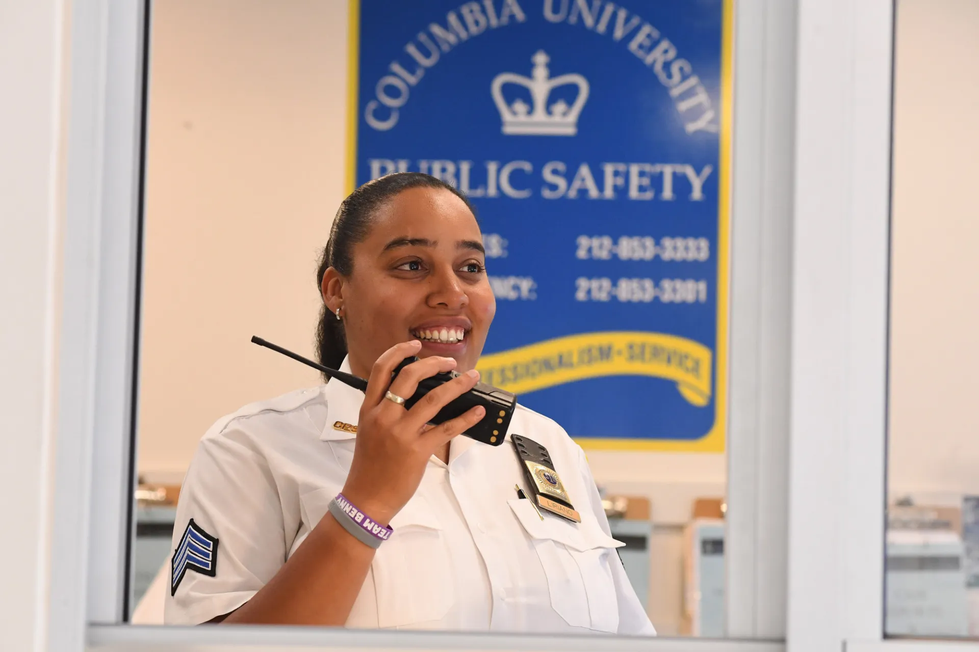 A Public Safety employee uses the radio to speak to someone. Behind her, a blue sign reads, "Columbia University Public Safety."