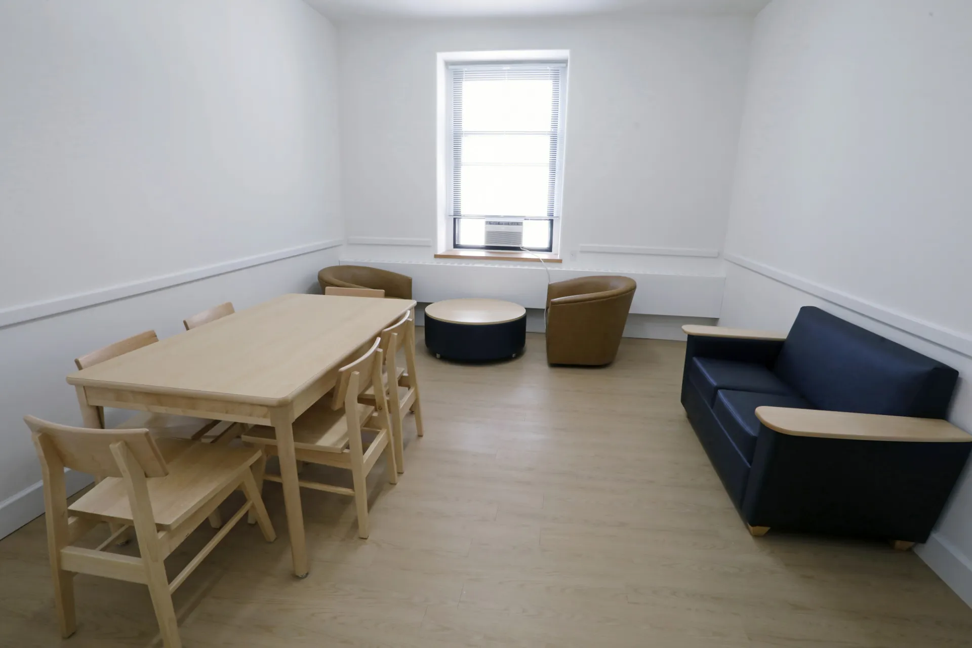 A refreshed lounge in Hogan Hall with a light wood table, a blue couch, two tan chairs and a coffee table.
